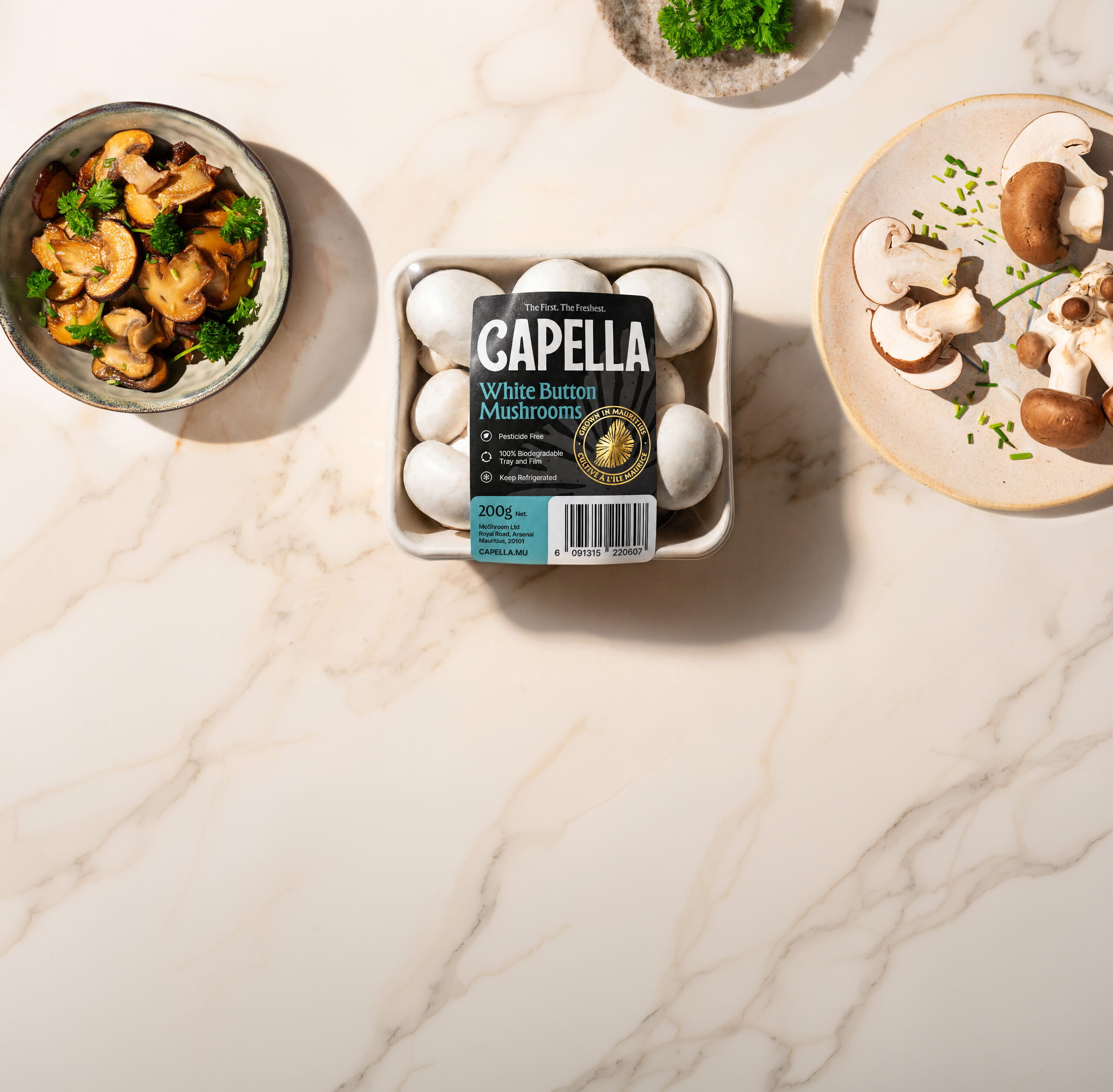 Capella White Button Mushrooms in a tray on a marble surface, surrounded by cooked and sliced mushrooms